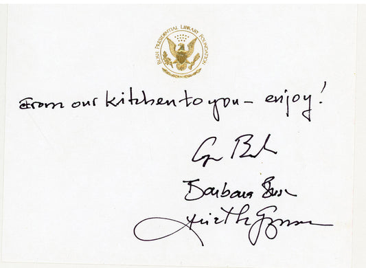 A white card with a raised presidential gold seal at the top, a handwritten message reading 'from our kitchen to you - enjoy!', and three signatures including George Bush, Barbara Bush and Ariel De Guzman.