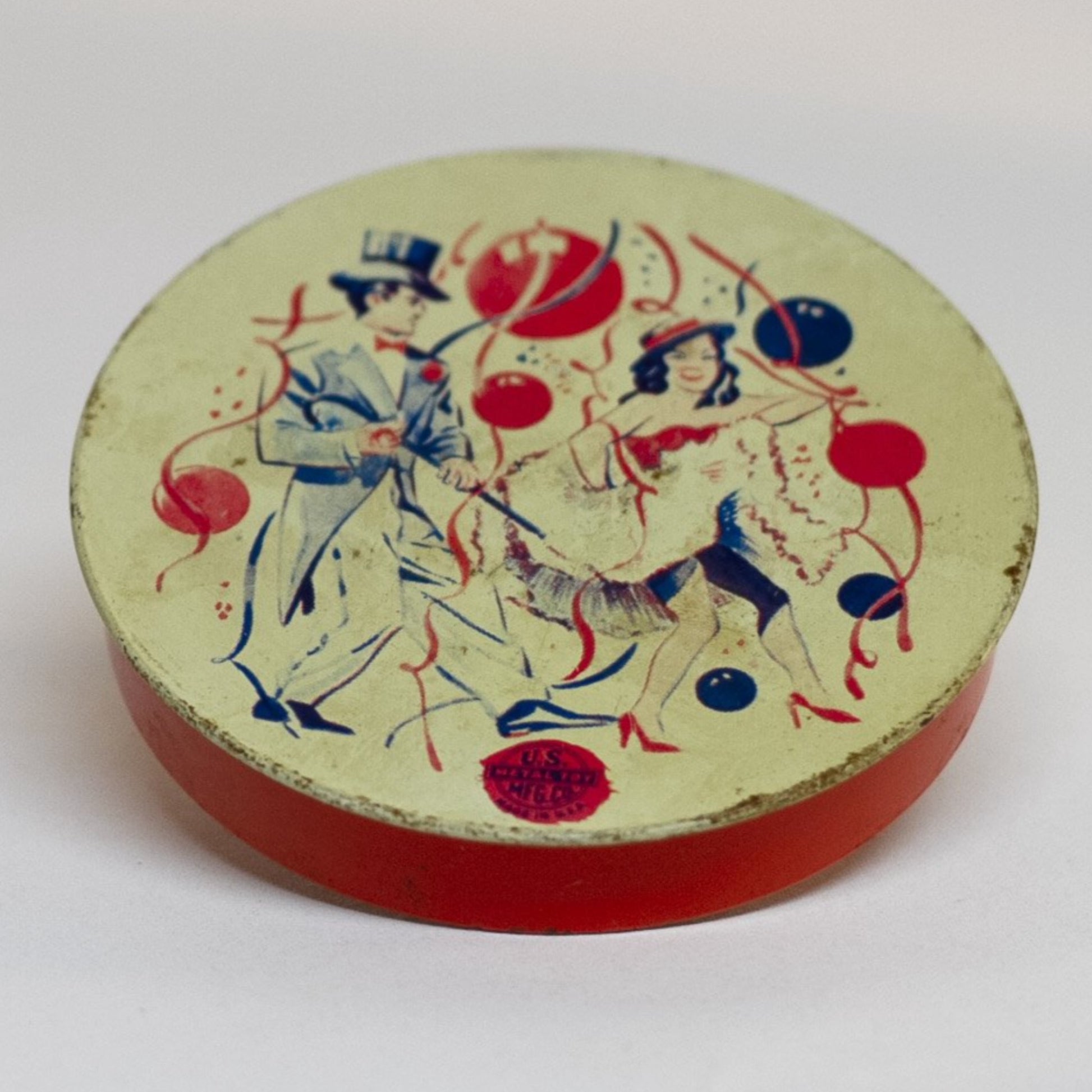 This round TIN-LITHOGRAPH RATCHET-STYLE NEW YEARS EVE NOISEMAKER made by US Metal Toys from the late 1940s to 1950s depicts a couple, a man dressed in a top hat and tailcoat and a woman in cha-cha costume dancing. The light-colored background is filled with red and blue balloons and ribbon streamers.