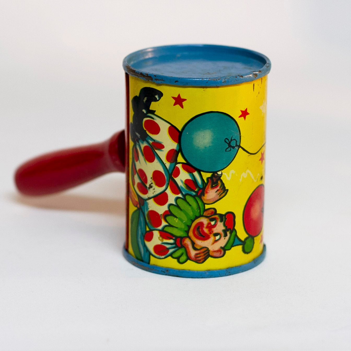 This is an older style tin LITHOGRAPH CLOWN NOISEMAKER that likely dates to the 1930s or 1940s is in the shape of a small can. The lithograph depicts two clowns, one dressed in a white and red polka-dot clown suit and the other is "toothless" and dressed in blue plaid. Scene background in yellow. A red wooden handle is