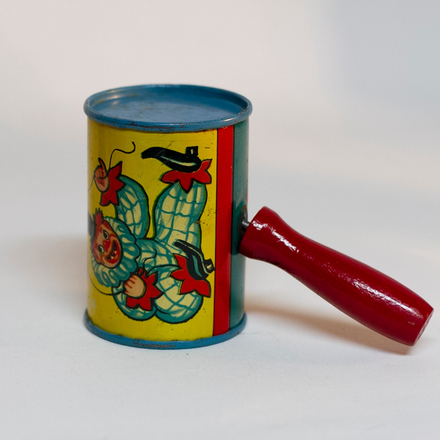 This is an older style tin LITHOGRAPH CLOWN NOISEMAKER that likely dates to the 1930s or 1940s is in the shape of a small can. The lithograph depicts two clowns, one dressed in a white and red polka-dot clown suit and the other is "toothless" and dressed in blue plaid. Scene background in yellow. A red wooden handle is