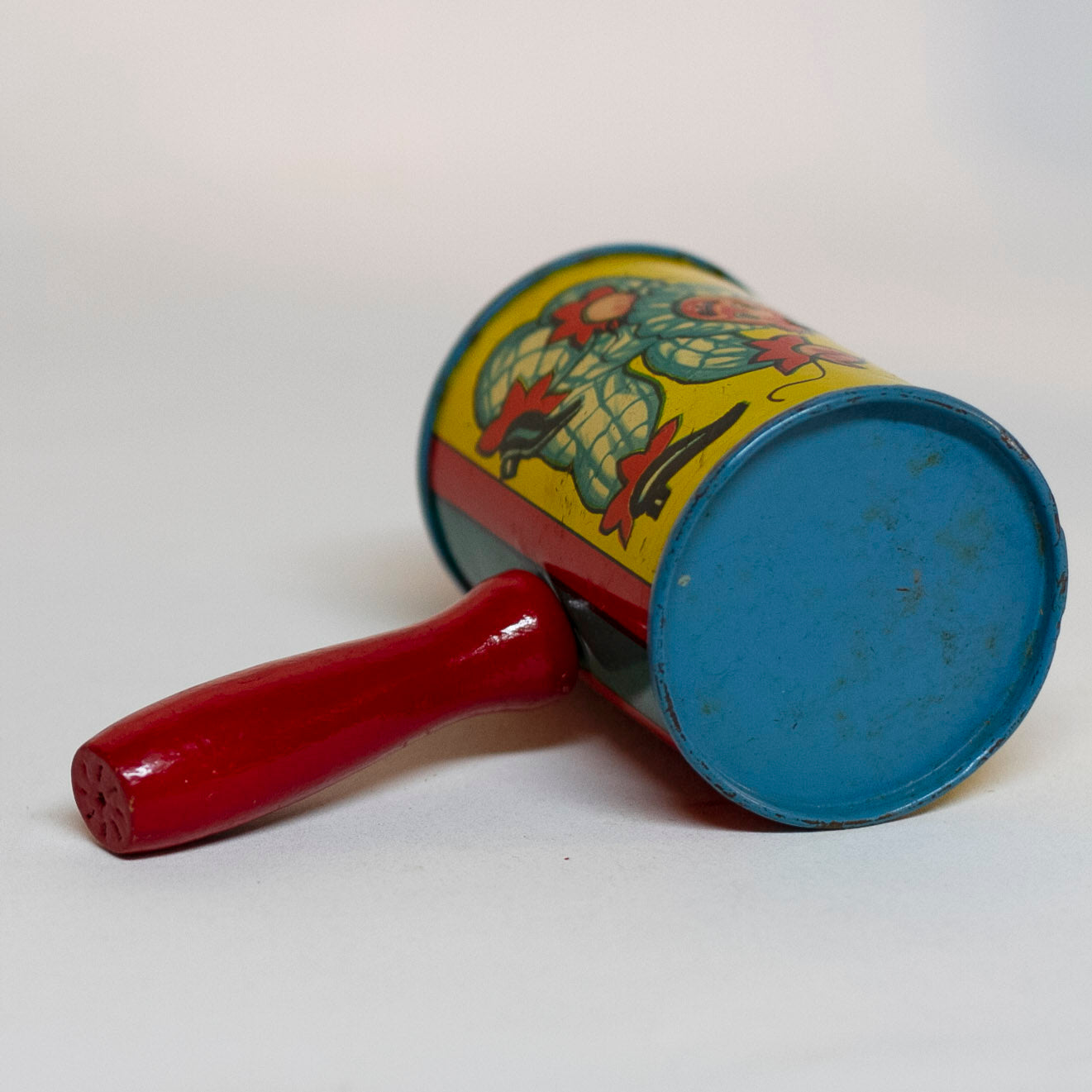 This is an older style tin LITHOGRAPH CLOWN NOISEMAKER that likely dates to the 1930s or 1940s is in the shape of a small can. The lithograph depicts two clowns, one dressed in a white and red polka-dot clown suit and the other is "toothless" and dressed in blue plaid. Scene background in yellow. A red wooden handle is