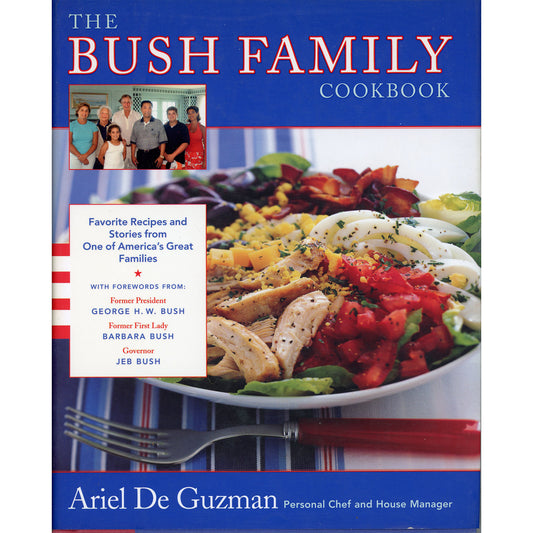 The Bush Family Cookbook by Ariel De Guzman, featuring a blue cover with the title in red and white, a family photo, a large prepared Cobb Salad on a plate, and a fork at the bottom.