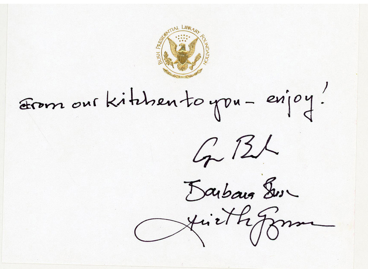 A white card with a raised presidential gold seal at the top, a handwritten message reading 'from our kitchen to you - enjoy!', and three signatures including George Bush, Barbara Bush and Ariel De Guzman.