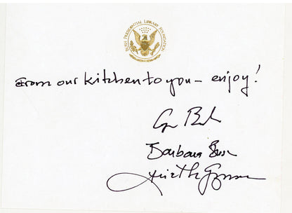 A white card with a raised presidential gold seal at the top, a handwritten message reading 'from our kitchen to you - enjoy!', and three signatures including George Bush, Barbara Bush and Ariel De Guzman.