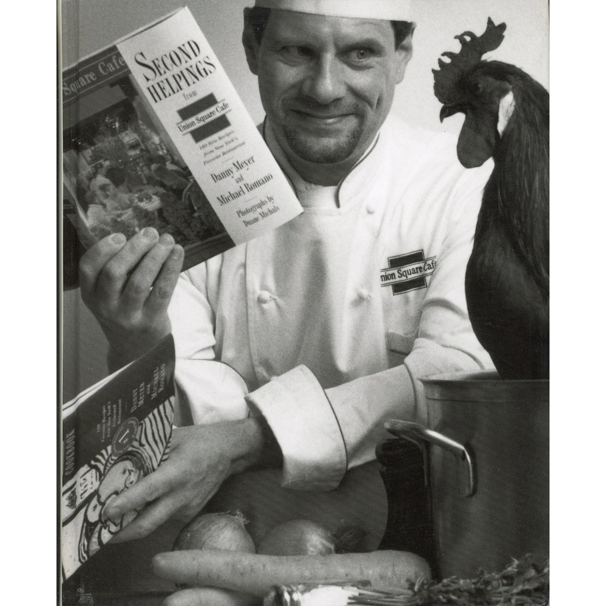 A black-and-white photo of a Danny Meyer in a white uniform holding a cookbook titled 'Second Helpings'  with a rooster standing beside a pot.
