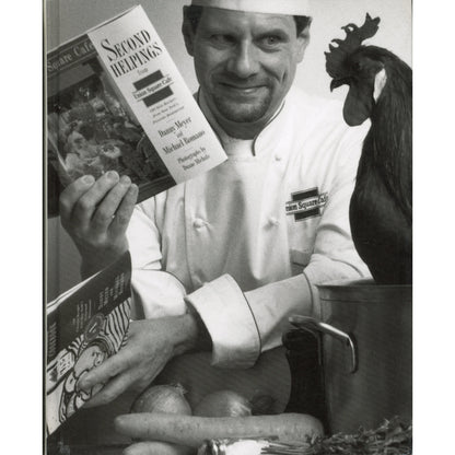 A black-and-white photo of a Danny Meyer in a white uniform holding a cookbook titled 'Second Helpings'  with a rooster standing beside a pot.