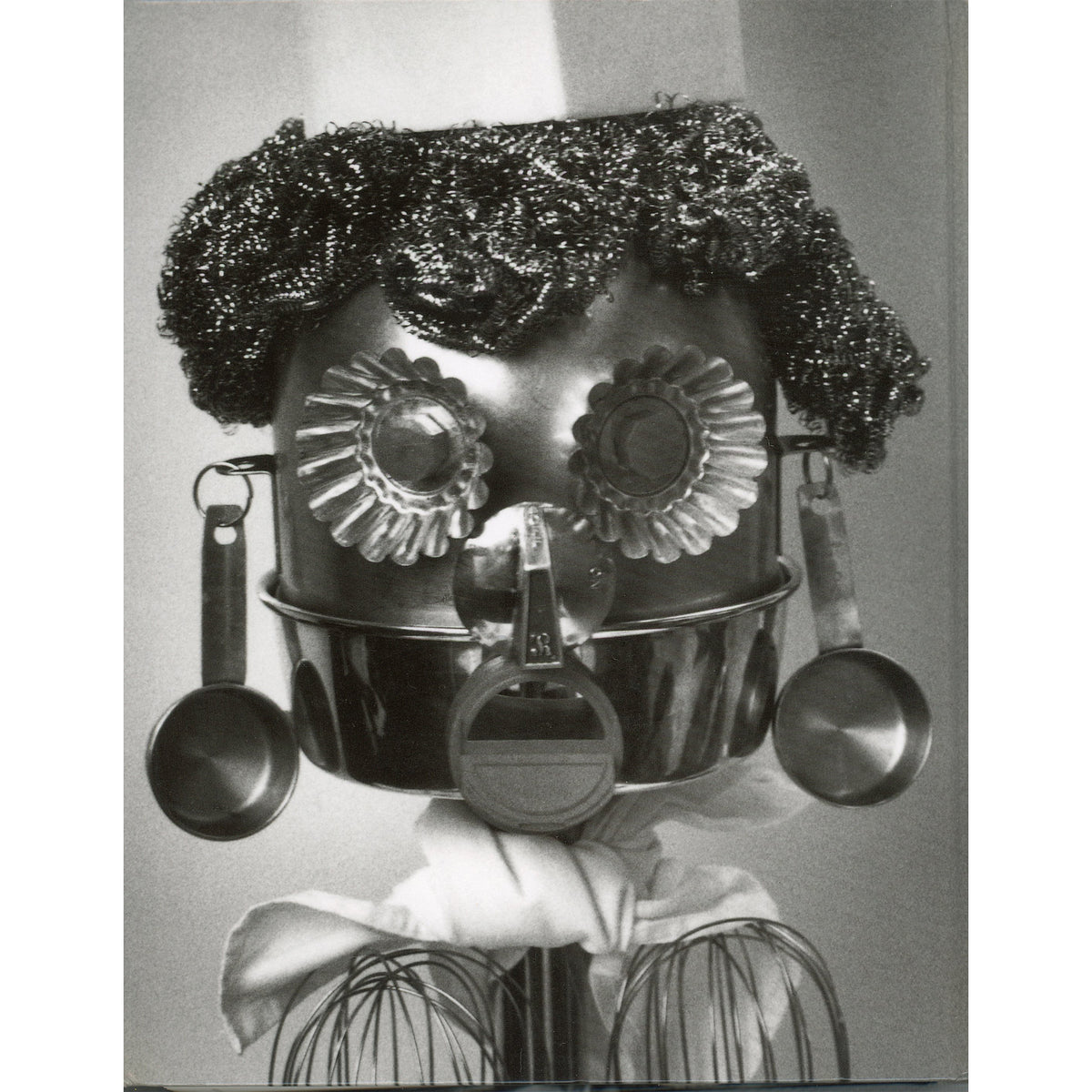 A black and white photograph on the back cover of the cookbook, Second Helpings from Union Square Cafe, of a whimsical, handmade robot or doll made from kitchen utensils, featuring a metal pot for a head with textured hair, flower-shaped eyes, hanging measuring cups, and wire whiskers.