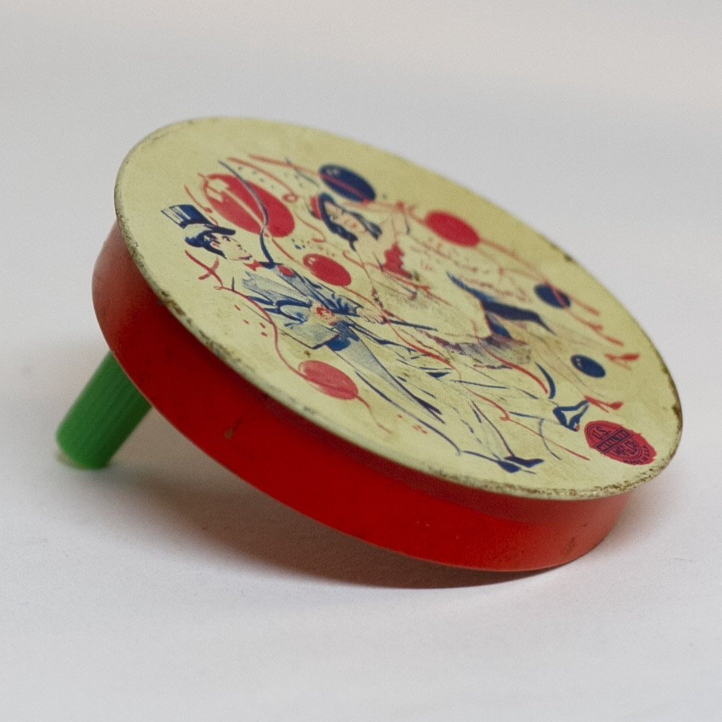 This round TIN-LITHOGRAPH RATCHET-STYLE NEW YEARS EVE NOISEMAKER made by US Metal Toys from the late 1940s to 1950s depicts a couple, a man dressed in a top hat and tailcoat and a woman in cha-cha costume dancing. The light-colored background is filled with red and blue balloons and ribbon streamers. 