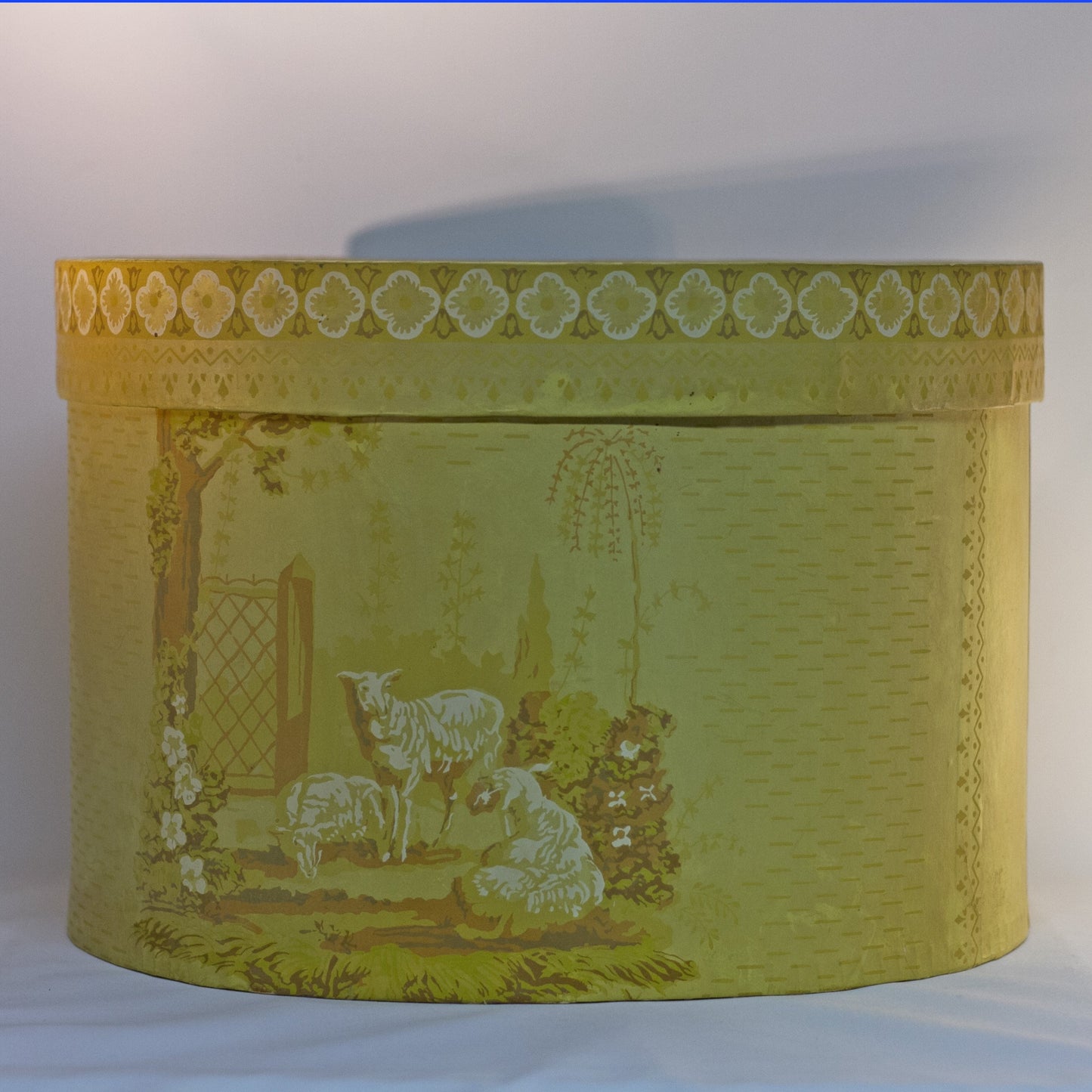 Reproduction of a "BRIDE'S BOX" made from slightly oval shaped large hat box covered in vintage yellow wallpaper featuring lambs and a Colonial girl. Inside and bottom papered with pages from the Maine Antique Digest, September 1977. Measures about 10" high by 15" in length by 13" wide.